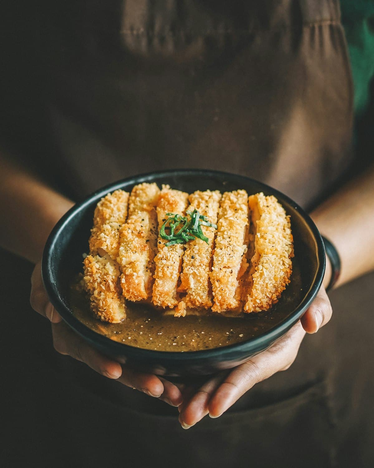 Krokante, gepaneerde kipreepjes op een bord met dikke Japanse currysaus, gegarneerd met lente-ui.